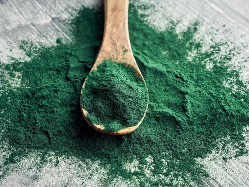 Spirulina powder in a wooden spoon on a textured gray surface.